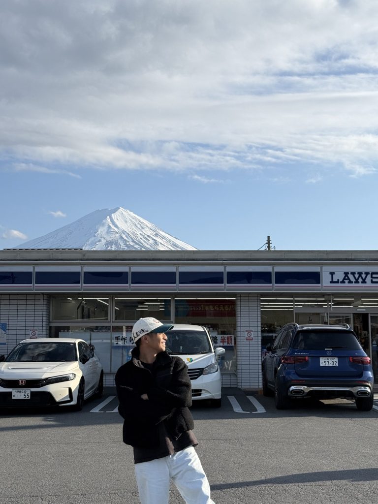 Man in a cap and white pants standing in front of the famous Lawson convenience store with snowcapped Mt. Fuji rising behind it