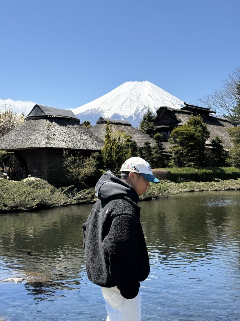 Man looking down at a clear pond at Oshino Hakkai, with thatched-roof houses and snowcapped Mt. Fuji behind