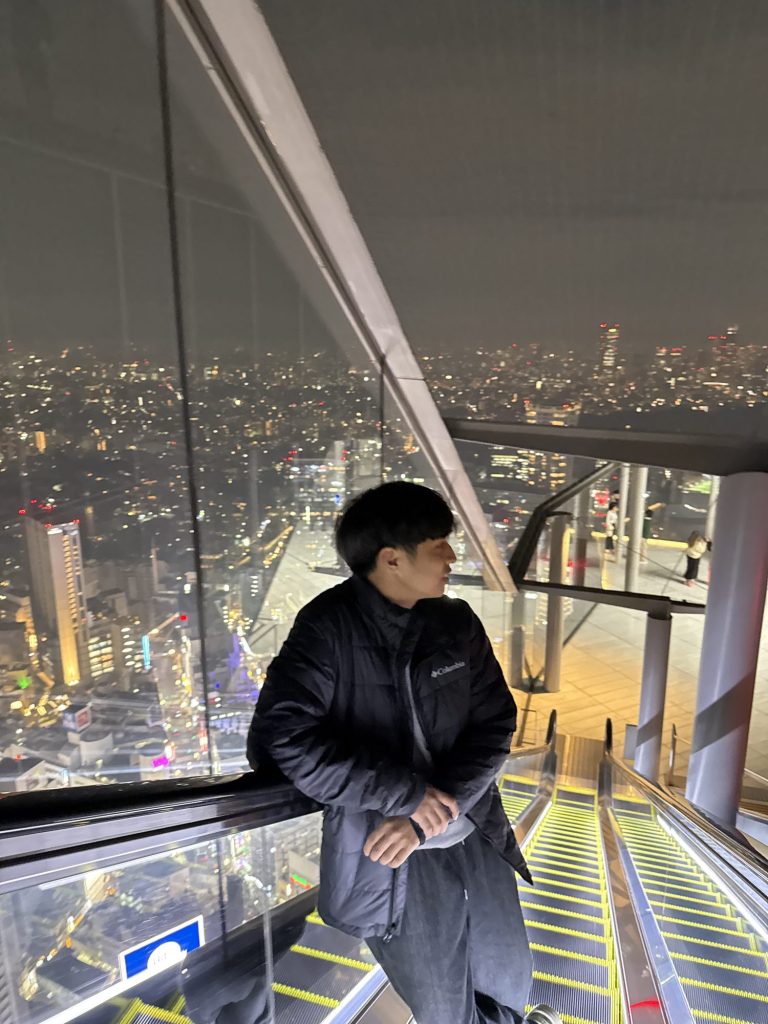 Man leaning on the glass at the top of the Shibuya Sky escalator with the city lights of Tokyo spread out below