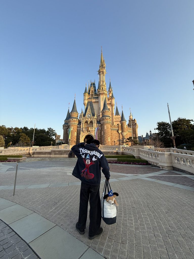 Man in a Disneyland jacket with a Duffy tote standing in front of Cinderella Castle under a clear blue sky