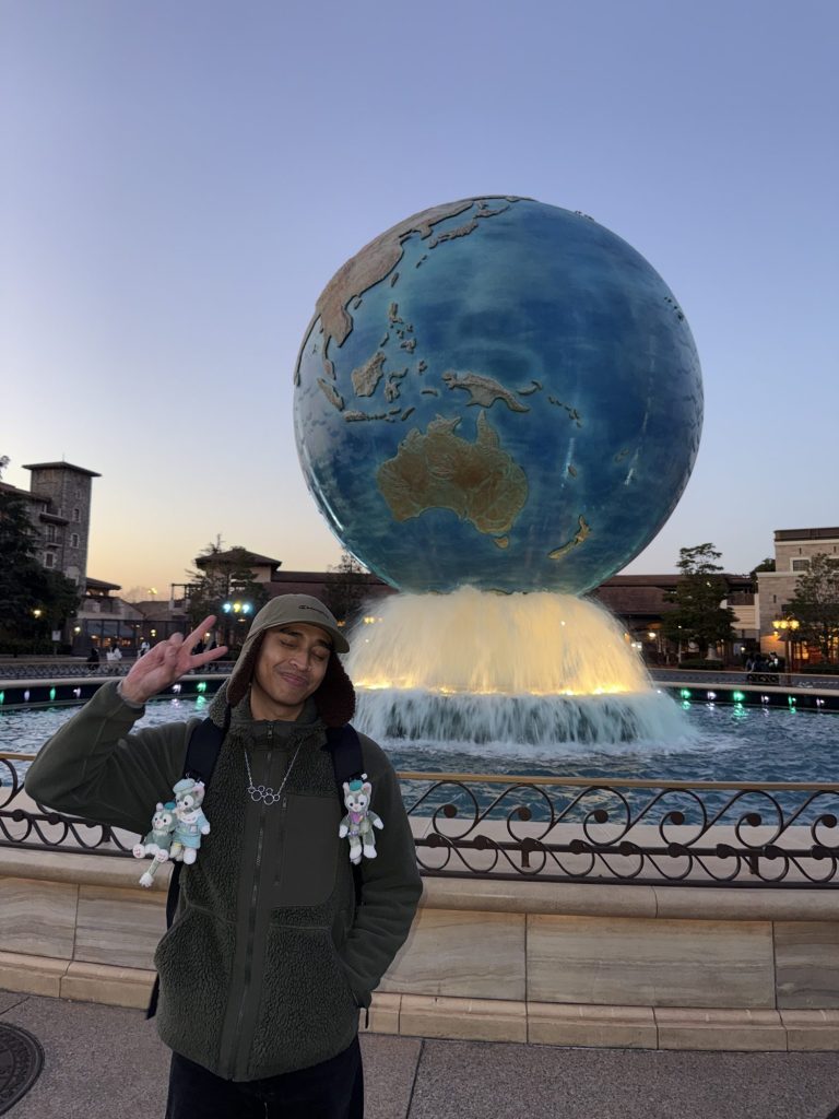Man throwing a peace sign in front of the giant AquaSphere globe fountain at the Tokyo DisneySea entrance at dusk