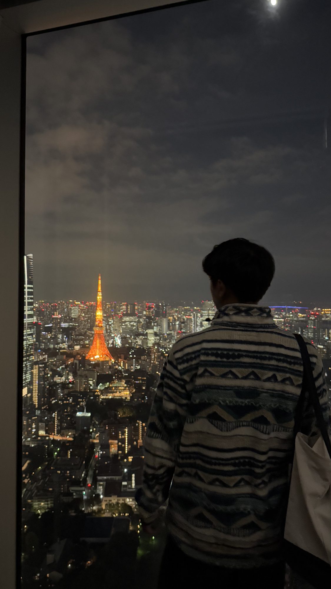 Man in a patterned jacket looking out a floor-to-ceiling window at a lit-up Tokyo Tower and the city at night from Roppongi Hills